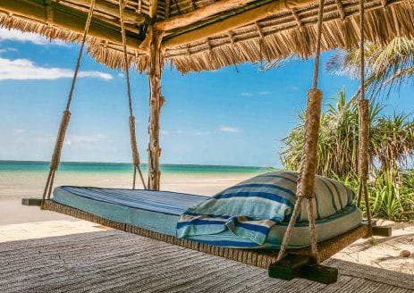 Hanging bed with blue-striped linens under a thatched roof, overlooking a sandy beach and turquoise ocean. Tropical plants are nearby, and its a sunny day with a few clouds in the sky.