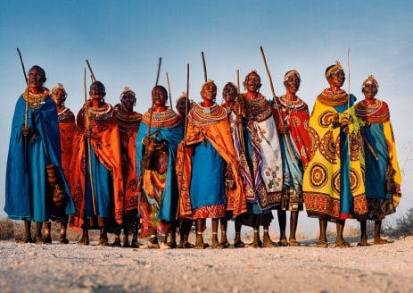 A group of people in colorful traditional attire stand together outdoors. They wear vibrant robes and intricate beadwork, and most hold sticks. The background includes a blue sky and a rocky landscape.